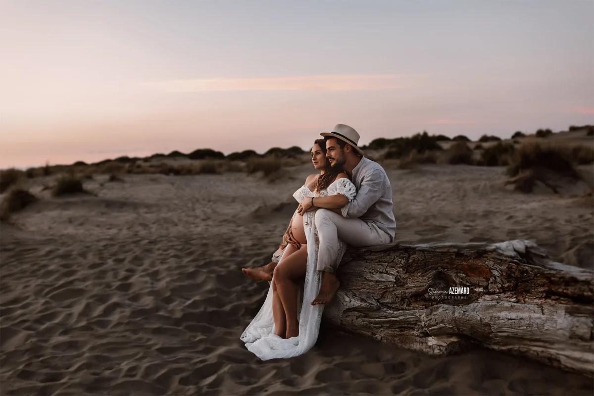 Photo grossesse couple assis sur tronc bois dans les dunes au coucher de soleil, photographe maternité Montpellier Stéphanie Azémard