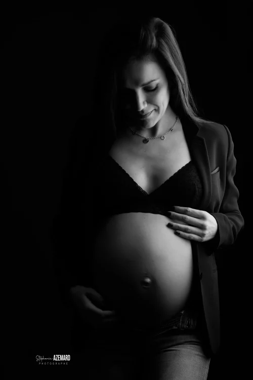 © Stéphanie Azémard — Photographe maternité Montpellier Photo grossesse future maman souriante en blazer ouvert tenant son ventre arrondi en noir et blanc studio fond noir, photographe maternité Montpellier Stéphanie Azémard