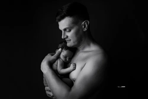 © Stéphanie Azémard — Photographe maternité Montpellier Photo papa souriant tenant tendrement son nouveau-né endormi contre sa poitrine en noir et blanc studio fond noir, photographe maternité Montpellier Stéphanie Azémard