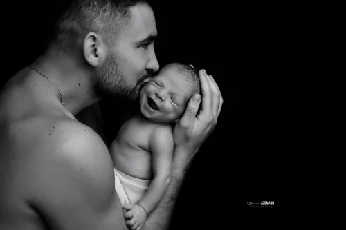 © Stéphanie Azémard — Photographe maternité Montpellier Photo nouveau-né souriant pendant que son papa lui fait un bisou sur la joue en noir et blanc studio fond noir, photographe maternité Montpellier Stéphanie Azémard